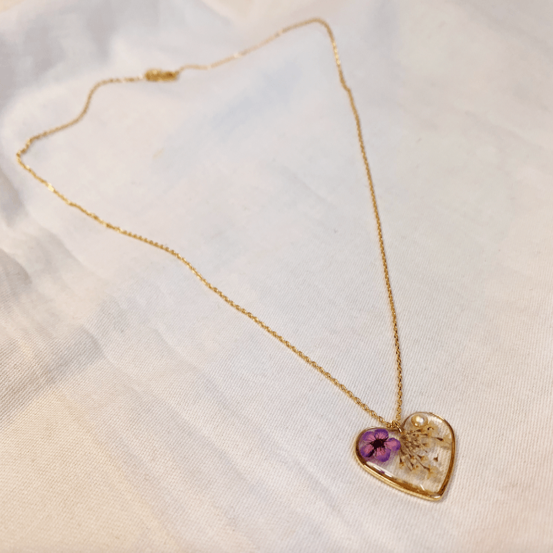 14ct Gold necklace with a heart pendent hanging from it. The pendent made form polished clear resin, encapsulating a dainty purple flower, a white flower and a pearl. The pendent rounded with  a gold plated frame 